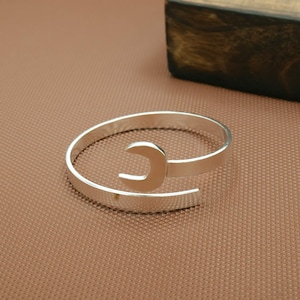 May include: A silver-toned cuff bracelet featuring a wrench design. The open-ended wrench is the focal point, set against a simple, polished band. The bracelet is displayed on a textured brown surface.