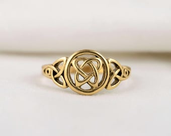 May include: A gold-toned ring with a Celtic knot design. The ring showcases a central circular motif with an elaborate knot, complemented by decorative details on the band. The ring is presented on a soft, white backdrop.