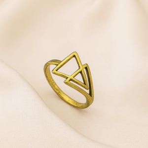 May include: A gold-coloured ring with a geometric design of two overlapping triangles. The ring has a simple band and a textured finish, set against a soft, off-white background. The ring's design is modern and minimalist.