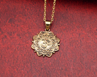 May include: Gold-coloured pendant necklace featuring a detailed Om symbol within a floral design. The pendant hangs from a delicate gold chain. The necklace is displayed against a red background.