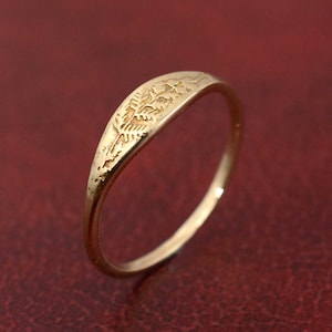 May include: A gold-colored ring with a detailed, textured design on the top surface. The ring has a smooth, polished band and a slightly curved shape. The design appears to be an abstract pattern or possibly a stylized leaf.