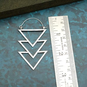 Op de afbeelding: Zilveren oorbel met een geometrisch ontwerp van drie gestapelde driehoeken. De oorbel is ongeveer 5 cm lang, gemeten met de liniaal in de afbeelding. De oorbel heeft een haaksluiting.