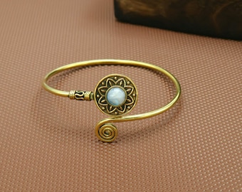 Puede incluir: Una pulsera dorada con una pieza central circular con un diseño floral. El centro del diseño floral presenta una piedra azul claro. La pulsera tiene un diseño en espiral en un extremo y un cierre adornado en el otro.