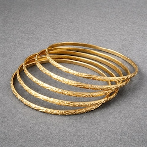 May include: Six gold bangles with a textured floral design. The bangles are stacked on a gray surface. The bangles are made of a shiny gold material.