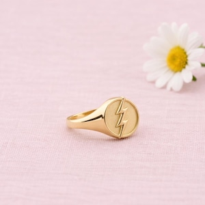 Op de afbeelding: Een goudkleurige ring met een bliksemontwerp op een cirkelvormig vlak. De ring staat tegen een zachtroze achtergrond met een madeliefbloem. De ring is een sieraad.