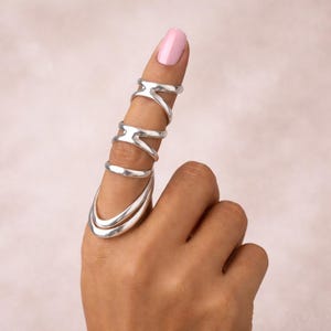 May include: A silver finger ring set. The set includes a ring that covers the entire finger, with three additional rings. The rings have a smooth, polished finish. The background is a soft pink.