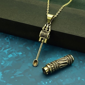 May include: A gold-coloured pendant necklace with an intricate design. The pendant includes a cylindrical element with a spoon-shaped attachment and a separate, matching cylindrical piece. The necklace is shown on a blue surface.