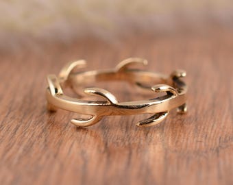 May include: A gold-colored ring with a delicate, twig-like design. The ring features multiple curved branches that wrap around the band. The ring is set against a wooden surface.
