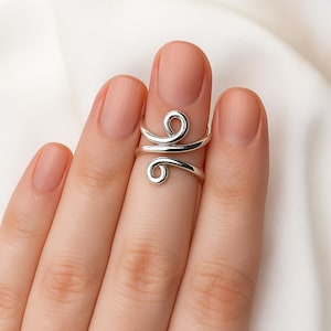 Può includere: Un anello regolabile color argento con un design a spirale. L'anello presenta due spirali, una in alto e una in basso, collegate da una fascia curva. L'anello è indossato su un dito.