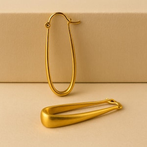 May include: Two gold-colored oval hoop earrings. One earring is upright, showing the clasp, while the other lies flat. The earrings have a smooth, matte finish and a simple, elegant design. The background is a neutral beige.
