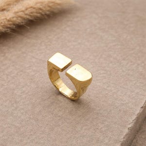 May include: A gold ring with a distinctive open design. The ring has two rectangular ends that don't meet, giving it a modern, minimalist look. It's shown on a neutral surface, emphasizing its polished finish. The ring's design is contemporary.