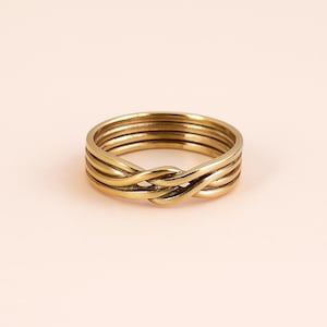 May include: A gold-colored ring with a woven design. The ring features multiple bands that intertwine to create a unique, textured appearance. The ring is made of a shiny, metallic material and is set against a soft, peach-colored background.