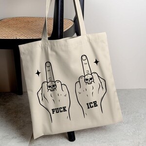 May include: Beige tote bag featuring a black graphic of two hands with middle fingers extended, each wearing a skull ring. The word "FUCK" is below one hand, and "ICE" is below the other. Black starbursts are on either side.
