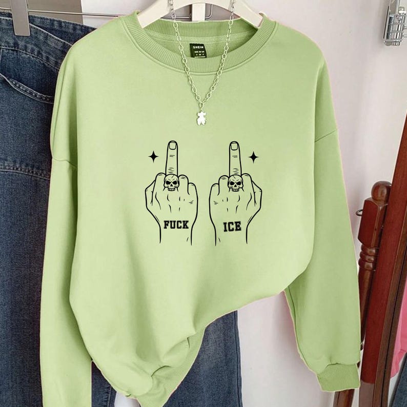 May include: Light green sweatshirt with a crew neck and long sleeves. The graphic features two hands with the middle fingers raised, each with a skull ring, and the words "FUCK" and "ICE" below. A silver necklace with a bear pendant is visible.