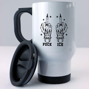May include: White travel mug with a black handle and lid. The mug features a black graphic of two skeleton hands giving the middle finger, with the words "FUCK ICE" below. The design includes stars and barbed wire details.