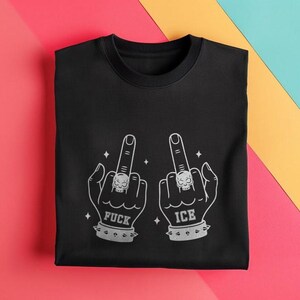 May include: Black crew neck t-shirt with a graphic design of two hands giving the middle finger. Each hand has a skull ring and a studded wristband. The word "FUCK" is printed under one hand, and "ICE" under the other.