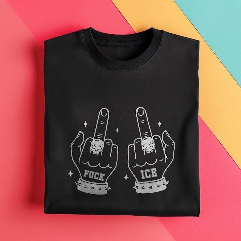 May include: Black crew neck t-shirt with a graphic design of two hands giving the middle finger. Each hand has a skull ring and a studded wristband. The word "FUCK" is printed under one hand, and "ICE" under the other.
