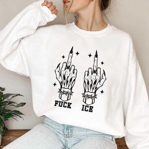 May include: White sweatshirt featuring a black graphic of two skeleton hands giving the middle finger. The hands are adorned with rings and surrounded by stars. The text "FUCK ICE" is printed below the hands.