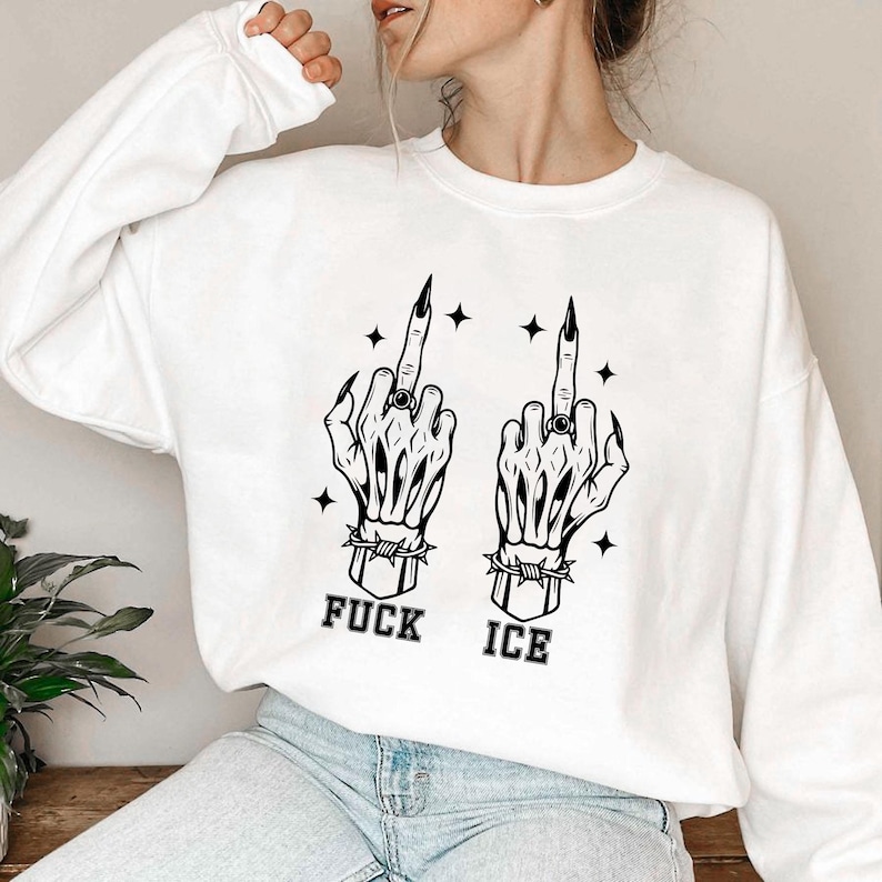 May include: White sweatshirt featuring a black graphic of two skeleton hands giving the middle finger. The hands are adorned with rings and surrounded by stars. The text "FUCK ICE" is printed below the hands.