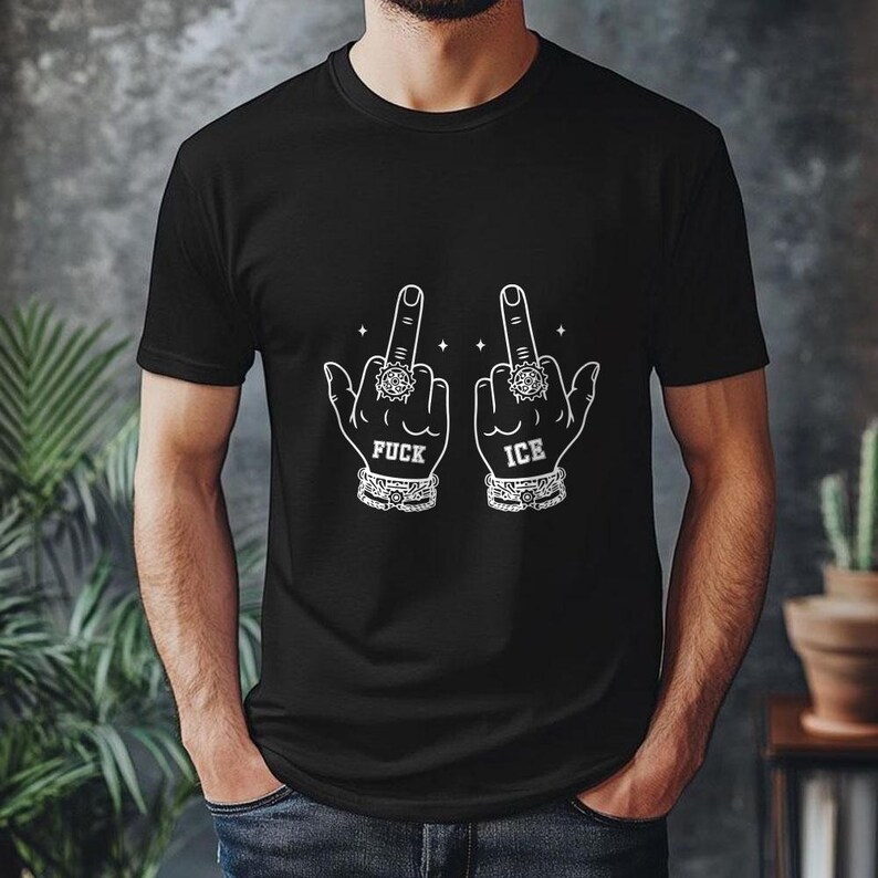May include: Black t-shirt with a white graphic of two hands displaying the middle finger. The text "FUCK ICE" is printed below the hands. The hands are adorned with rings and bracelets.