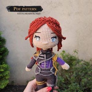Può includere: Una bambola all'uncinetto fatta a mano con capelli rossi, occhi blu e un abito dettagliato nei toni del grigio, viola e oro. La bambola è tenuta in mano e presenta il testo "PDF PATTERN" e "stitchesbysunky".