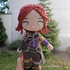 May include: A handmade crochet doll with red hair and blue eyes. The doll wears a detailed outfit in shades of brown, purple, and gold. The doll's hair is styled in a unique fashion, and the doll is approximately 8 inches tall.