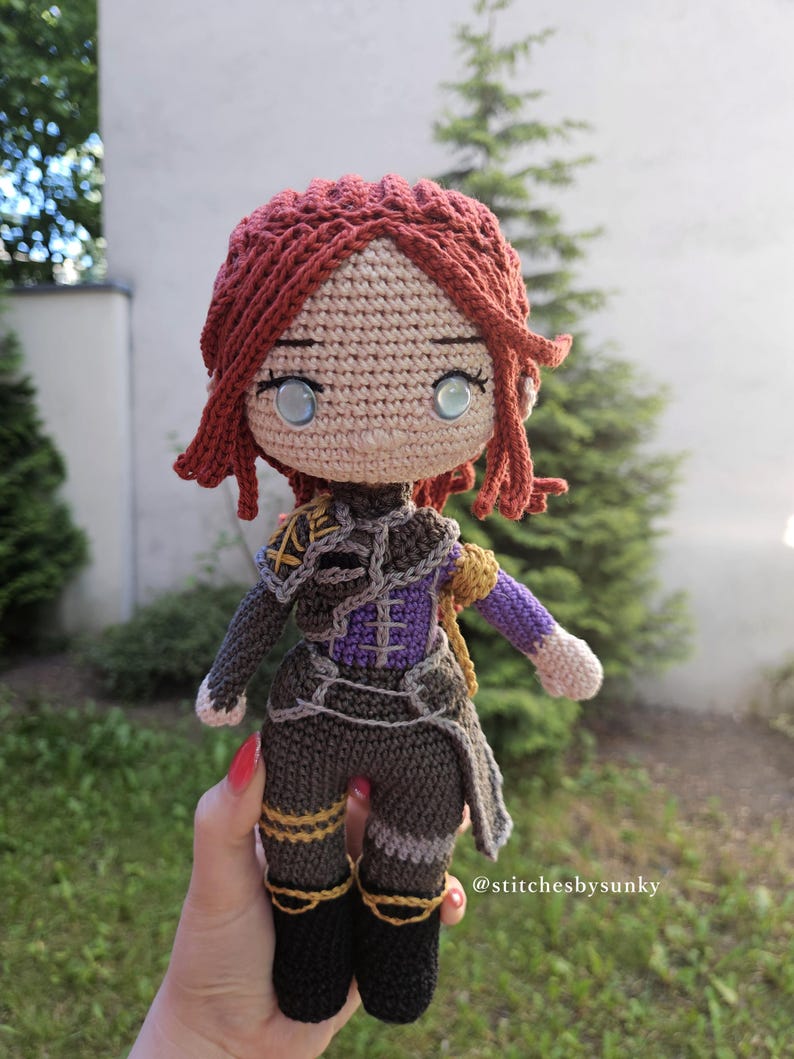 May include: A handmade crochet doll with red hair and blue eyes. The doll wears a detailed outfit in shades of brown, purple, and gold. The doll's hair is styled in a unique fashion, and the doll is approximately 8 inches tall.