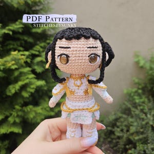 May include: A handmade crochet doll with black hair, brown eyes, and a white and gold outfit. The doll is held in a hand. The image includes the text "PDF PATTERN" and "STITCHESBYSUNKY".