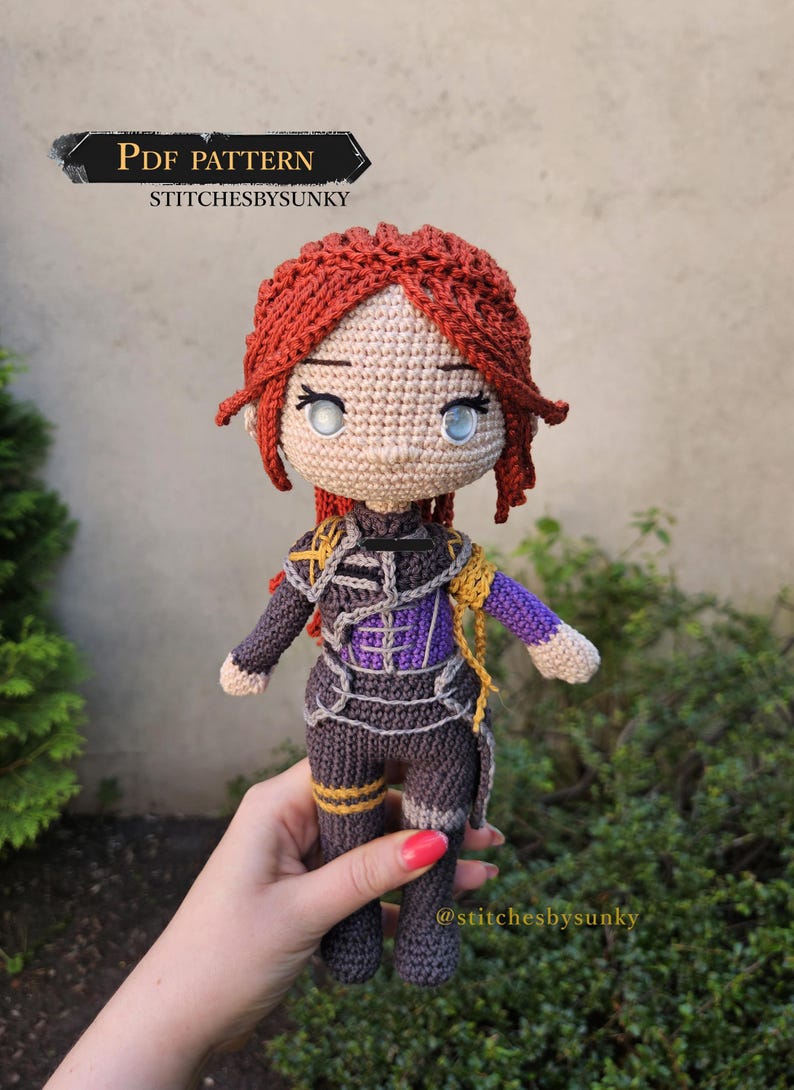 May include: A handmade crochet doll with red hair, blue eyes, and a detailed outfit in shades of gray, purple, and gold. The doll is held in a hand and features the text "PDF PATTERN" and "stitchesbysunky".