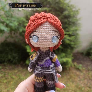 Puede incluir: Muñeca de ganchillo hecha a mano con cabello rojo, ojos azules y un atuendo detallado en tonos grises, morados y dorados. La muñeca sostiene una espada. El texto "PDF PATTERN" y "STITCHESBYSUNKY" son visibles.