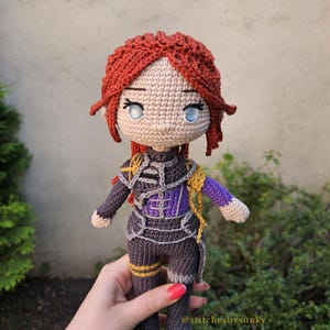 May include: Crocheted doll with red hair, blue eyes, and a detailed outfit in shades of gray, purple, and gold. The doll is handmade, with intricate stitching visible throughout the design. The doll is approximately 8 inches tall.