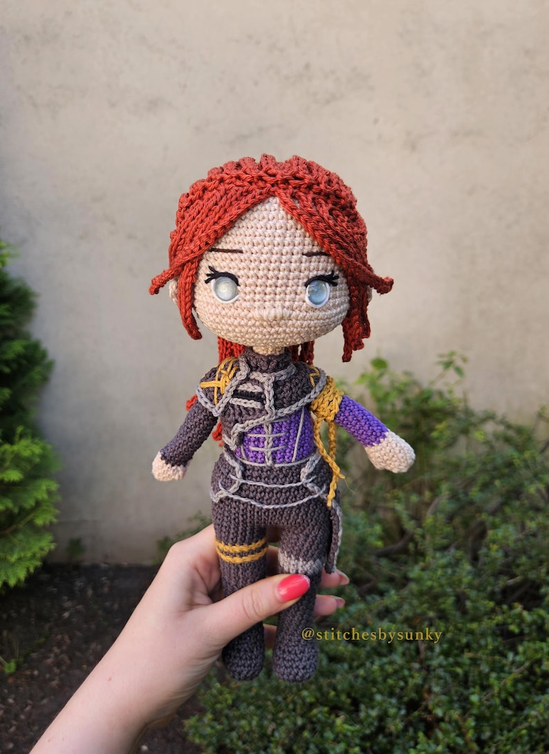 May include: Crocheted doll with red hair, blue eyes, and a detailed outfit in shades of gray, purple, and gold. The doll is handmade, with intricate stitching visible throughout the design. The doll is approximately 8 inches tall.