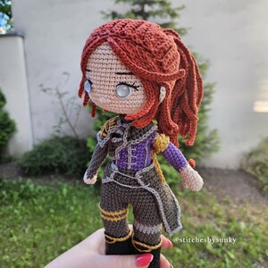 May include: A handmade crochet doll with red hair styled in a ponytail. The doll wears a detailed outfit in shades of gray, purple, and gold. The doll's eyes are light blue. The doll is held in a hand. The text "@stitchesbysunky" is visible.