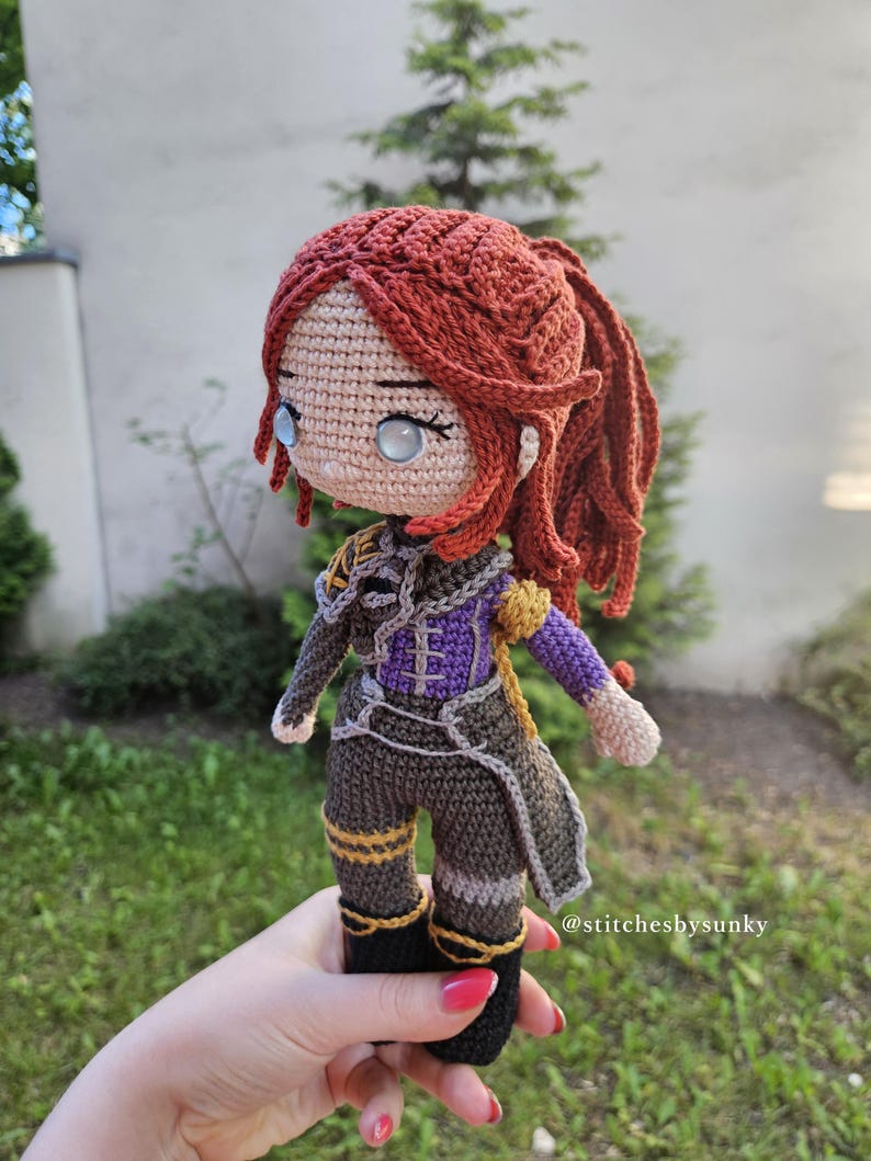 May include: A handmade crochet doll with red hair styled in a ponytail. The doll wears a detailed outfit in shades of gray, purple, and gold. The doll's eyes are light blue. The doll is held in a hand. The text "@stitchesbysunky" is visible.