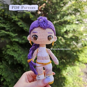 Puede incluir: Muñeca de ganchillo hecha a mano con peinado morado y detalles dorados. La muñeca lleva un atuendo blanco y amarillo. El texto "PDF PATTERN" y "stitchesbysunky" son visibles en la imagen. La muñeca está sujeta en una mano.