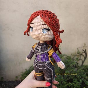 May include: A handmade crochet doll with red hair and blue eyes. The doll is wearing a dark gray and purple outfit with gold accents. The doll is being held by a person. The text "@stitchesbysunky" is visible.