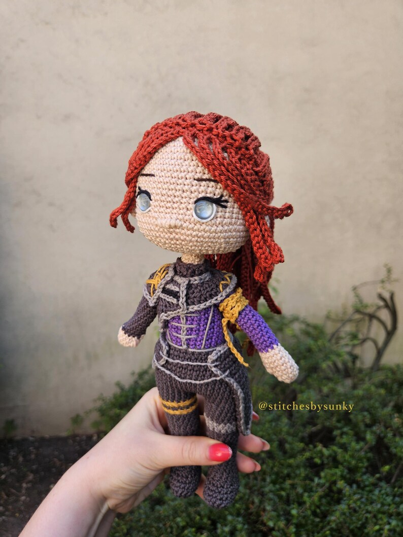 May include: A handmade crochet doll with red hair and blue eyes. The doll is wearing a dark gray and purple outfit with gold accents. The doll is being held by a person. The text "@stitchesbysunky" is visible.