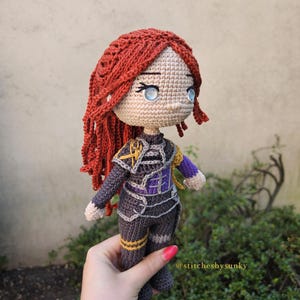 May include: A handmade crochet doll with red hair and blue eyes. The doll is wearing a dark gray outfit with purple and gold accents. The doll is held in a hand, with the text "@stitchesbysunky" visible.