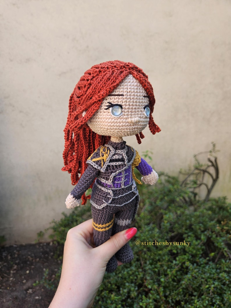 May include: A handmade crochet doll with red hair and blue eyes. The doll is wearing a dark gray outfit with purple and gold accents. The doll is held in a hand, with the text "@stitchesbysunky" visible.