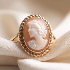 Puede incluir: Un anillo de oro con un camafeo ovalado que presenta el perfil tallado en blanco de una persona sobre un fondo color melocotón. El camafeo está enmarcado por un borde de oro trenzado tipo cuerda y se asienta sobre una banda de oro fina.