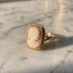 May include: A gold ring with a cameo portrait in a rectangular frame. The cameo is a light peach colour, and the frame has a rope-like texture. The ring is set against a marble surface.