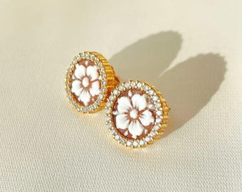 May include: A pair of gold-toned stud earrings. Each earring features a white flower design set against a brown background, surrounded by a ring of small, clear crystals. The earrings are round and have a classic, elegant style.