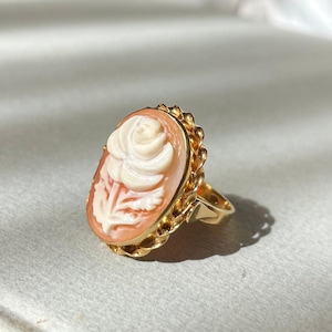Op de afbeelding: Een gouden ring met een ovale camee met een gesneden rozenontwerp. De camee is perzik van kleur met een witte roos en steel. De ring heeft een getextureerde gouden band en zetting.