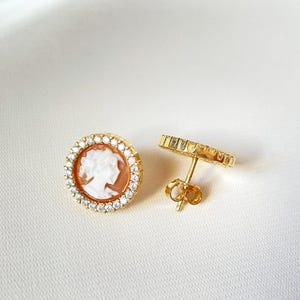 May include: Gold-tone stud earrings with a cameo design. Each earring features a round, orange and white cameo, encircled by small, clear crystals. The earrings are displayed on a white background.