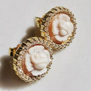 May include: A pair of gold-coloured stud earrings. Each earring features a carved white rose design set within a circular frame of clear, sparkling crystals. The rose is set against a light peach-coloured background.