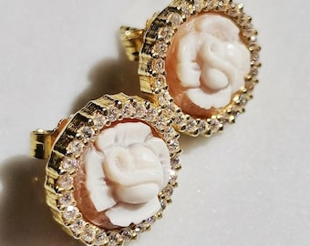 May include: A pair of gold-toned stud earrings. Each earring features a carved white rose design set within a circular frame of clear, sparkling crystals. The rose is set against a light peach-colored background.
