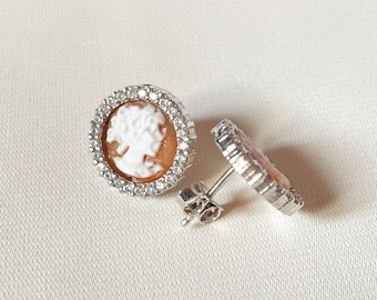 May include: A pair of silver stud earrings featuring a cameo design. Each earring has a round, bezel-set cameo with a white profile against a peach background, surrounded by a halo of small, clear crystals. The earrings are displayed on a white surface.