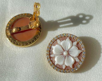 May include: A pair of clip-on earrings with a floral design. Each earring features a round, peach-colored cameo with a white flower carving, surrounded by a gold-colored frame and small, clear gemstones. The clip-on mechanism is visible on one earring.