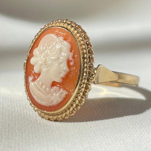 Puede incluir: Anillo de oro con un camafeo ovalado con el perfil de una mujer en crema sobre un fondo naranja. El anillo tiene un borde retorcido en forma de cuerda y una banda sencilla.