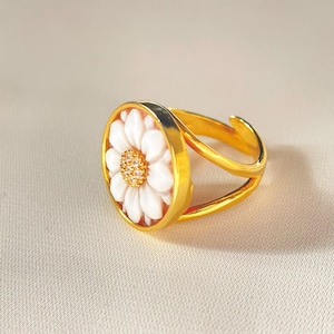 Puede incluir: Anillo dorado con un diseño de flor blanca y un centro brillante. El anillo tiene una banda dividida y una flor redonda engastada en bisel. Los pétalos de la flor están tallados y el centro está adornado con pequeñas piedras transparentes.